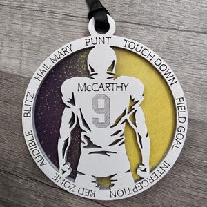 May include: A white wooden ornament shaped like a football player with the name "McCarthy" and the number "9" on the back. The ornament is decorated with purple and yellow glitter and has the words "Hail Mary", "Punt", "Touch Down", "Field Goal", "Interception", "Red Zone", and "Audible Blitz" around the edge.