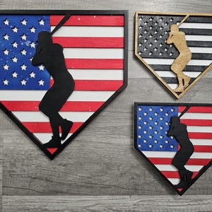 May include: Three wooden home plate signs with an American flag design and a silhouette of a baseball player. The flags are red, white, and blue with a distressed look. The home plates are black.
