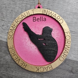 May include: A pink and gold wooden ornament with a silhouette of a person doing a back handspring. The ornament has the words "Back Forward Reverse Somersault Inward" written around the edge and the name "Bella" in the center.