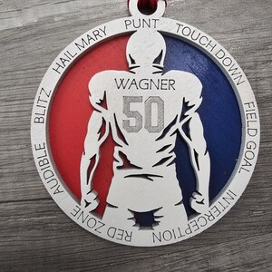 May include: A wooden ornament shaped like a football player with the words "PUNT", "TOUCH DOWN", "FIELD GOAL", "INTERCEPTION", "AUDIBLE BLITZ", "HAIL MARY", and "RED ZONE" around the edge. The player has the name "WAGNER" and the number "50" on the back. The ornament is painted red, white, and blue.