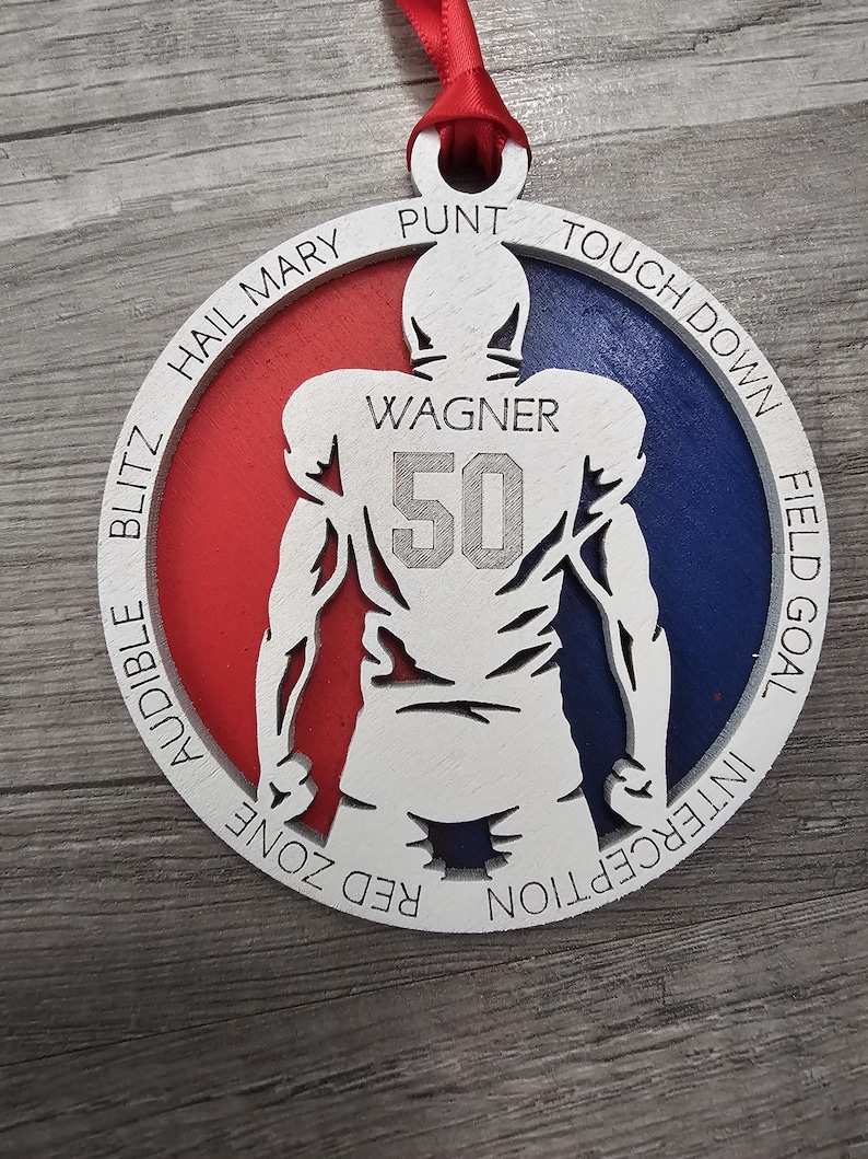 May include: A wooden ornament shaped like a football player with the words "PUNT", "TOUCH DOWN", "FIELD GOAL", "INTERCEPTION", "AUDIBLE BLITZ", "HAIL MARY", and "RED ZONE" around the edge. The player has the name "WAGNER" and the number "50" on the back. The ornament is painted red, white, and blue.