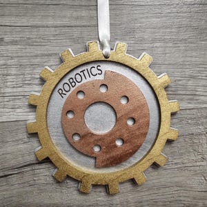 May include: A gear-shaped ornament featuring a gold outer rim, a silver middle layer, and a copper-colored center disc with circular cutouts. The word ROBOTICS is printed in black on the silver section. A white ribbon is attached to the top for hanging.