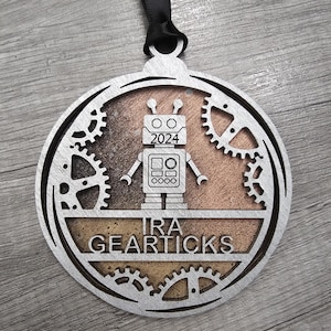 May include: A circular silver-toned ornament featuring a central robot figure with the year 2024 on its chest. Below the robot, the text IRA GEARTICKS is displayed. The design includes gear motifs and a textured bronze-colored background with a black ribbon.