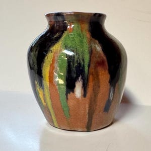 May include: A small ceramic vase with a rounded body and a short neck. The surface features a glossy glaze with vertical drips of black, forest green, bright yellow, and burnt orange over a textured, earthy brown base color.