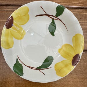 Puede incluir: Un cuenco redondo de cerámica con base blanca y un diseño floral pintado a mano. El patrón consta de dos grandes flores amarillas con centros de color marrón oscuro, unidas por tallos finos marrones y varias hojas verdes en el borde.