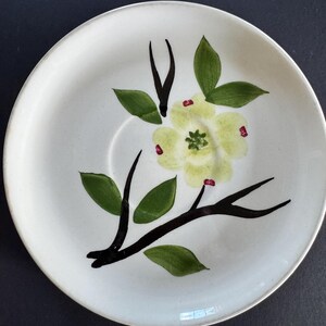 Op de afbeelding: Een rond, gebroken wit keramieken schoteltje met een handgeschilderd ontwerp van een enkele lichtgele bloem met een groen hart. Het bloemmotief bevat donkerbruine takken en diverse groene bladeren, met kleine rode accenten op de bloemblaadjes.