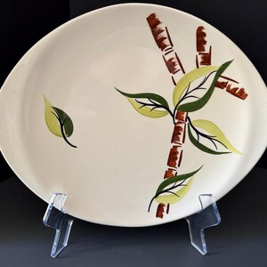 May include: An oval-shaped off-white ceramic platter featuring a hand-painted design of brown bamboo stalks and green leaves. The platter is supported by a clear acrylic stand, showcasing the botanical motif across the smooth, glossy surface.