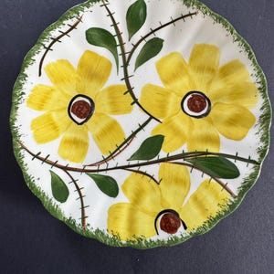 May include: A round ceramic plate with a scalloped edge featuring a hand-painted design of three large yellow flowers with brown centers. The floral pattern includes green leaves and thin brown stems with small cross-hatch markings. The rim is green.
