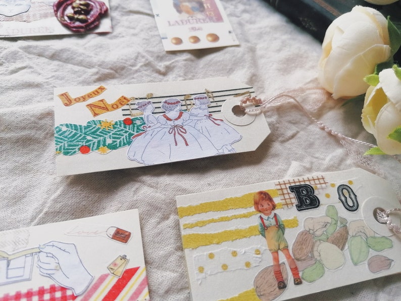 May include: A set of four Christmas-themed paper tags with various designs. One tag features a white background with three girls in white dresses and red ribbons, with the text "Joyeux Noel". Another tag has a yellow background with a girl in a red and white dress, with the text "BO" in black letters. The other two tags have a white background with various patterns and designs.