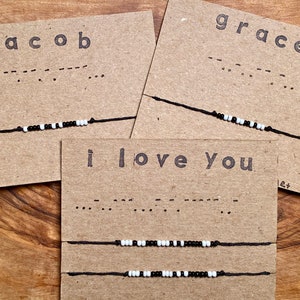 May include: Handmade Morse code bracelets on kraft paper cards. The cards display names and phrases like "Jacob", "Grace", and "I love you" in printed text and Morse code. The bracelets feature black and white beads on a black cord.