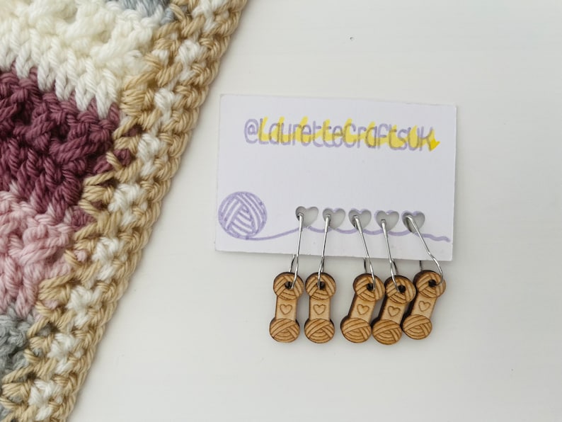 May include: A set of six wooden yarn stitch markers with a heart and yarn spool design. The markers are attached to silver rings and are displayed on a white card with the text "@laurettecraftsuk".