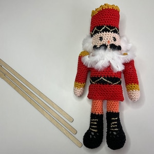 May include: A crocheted nutcracker doll with a red and gold hat, white beard, and black boots. The doll is standing on a white surface with three wooden sticks.