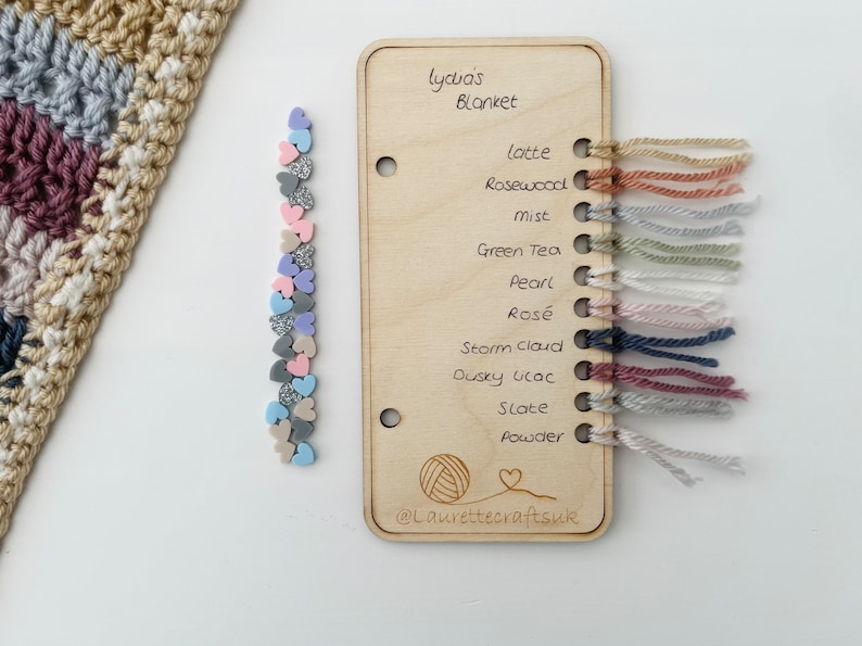 May include: A wooden card with the text "Lycia's Blanket" and a list of yarn colors with corresponding yarn samples. The colors are: Latte, Rosewood, Mist, Green Tea, Pearl, Ros&eacute;, Storm Cloud, Dusky Lilac, Slate, and Powder. The card also includes a small illustration of a ball of yarn and the text "@Laurettecraftsuk".