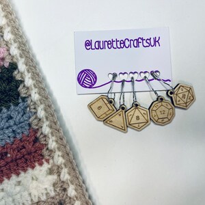 May include: Set of six wooden stitch markers shaped like dice with numbers 4, 6, 8, 12, and 20. The markers are attached to silver stitch holders. The stitch holders are hanging on a white card with the text "@LauretteCraftsUK" printed in purple.