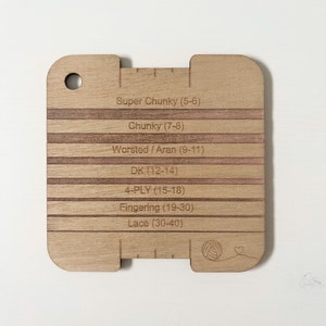 May include: A wooden gauge for measuring yarn thickness. The gauge has a scale with different yarn weights labeled: Super Chunky (5-6), Chunky (7-8), Worsted/Aran (9-11), DK (12-14), 4-PLY (15-18), Fingering (19-30), and Lace (30-40).