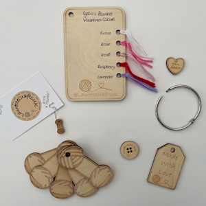 Peut inclure: Un ensemble de fournitures artisanales en bois pour la fabrication d'une couverture. L'ensemble comprend une carte en bois avec une liste de couleurs, des échantillons de fil, des pelotes de laine en bois, un bouton en bois, une étiquette en bois qui dit "Fabriqué avec amour" et un anneau métallique. La carte indique "Lydia's Blanket Valentines Colours" et liste les couleurs "Frost", "Rose", "Rose", "Raspberry" et "Lavender".