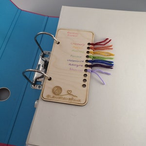 May include: A wooden card with a yarn sampler for different colors of yarn. The colors are: Rainbow Blanket, Cranberry, Pink, Mustard, Peridot, Ultramarine, Aubergine, Lavender. The card also has the text "@laurettecraftsuk" and a small illustration of a ball of yarn and a heart.