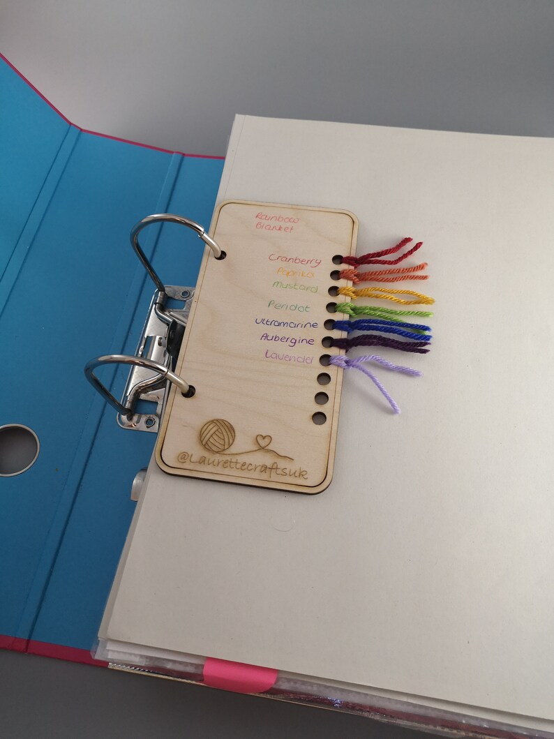 May include: A wooden card with a yarn sampler for different colors of yarn. The colors are: Rainbow Blanket, Cranberry, Pink, Mustard, Peridot, Ultramarine, Aubergine, Lavender. The card also has the text "@laurettecraftsuk" and a small illustration of a ball of yarn and a heart.