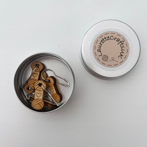 May include: A small silver tin with a label that says "Laurette Crafts UK" and a small heart. The tin contains several wooden yarn stitch markers with a small yarn ball and heart design.