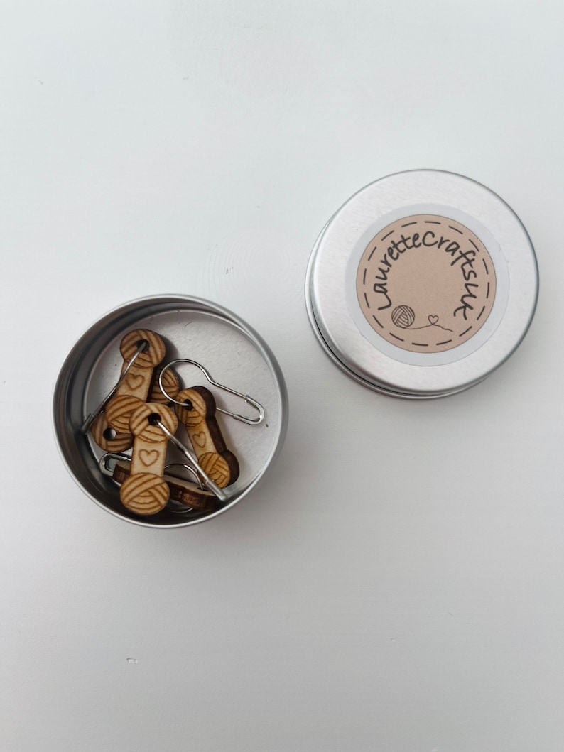 May include: A small silver tin with a label that says "Laurette Crafts UK" and a small heart. The tin contains several wooden yarn stitch markers with a small yarn ball and heart design.