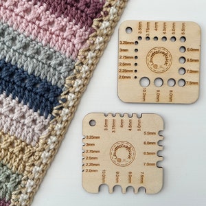 May include: Two wooden crochet hook gauge tools with different sizes of holes for measuring crochet hook sizes. The tools are labelled with measurements in millimetres. The tools are on a white surface.