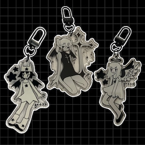 May include: Three black and white cartoon character keychains with silver key rings. The characters are a girl with wings, a girl with a cross, and a girl with a star on her head. Each keychain has the word "gross" printed on it.
