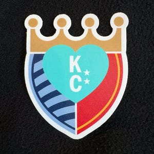 May include: A white heart-shaped shield with a blue and white striped section, a red section, and a gold crown on top. The heart is turquoise blue with the letters "K*C*" in white.