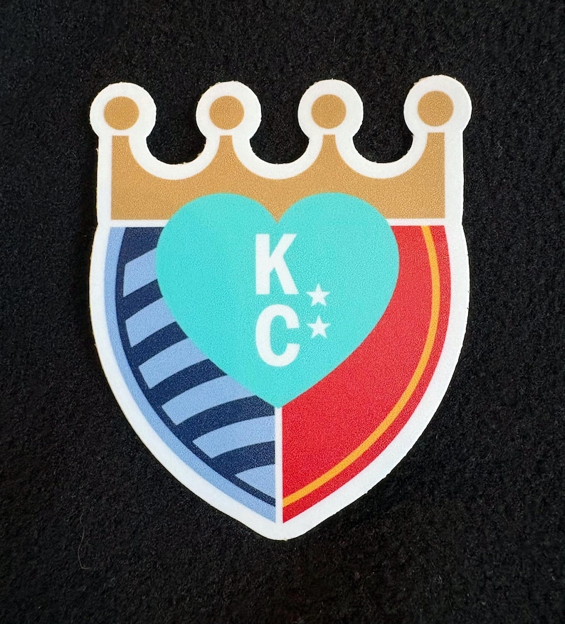 May include: A white heart-shaped shield with a blue and white striped section, a red section, and a gold crown on top. The heart is turquoise blue with the letters "K*C*" in white.