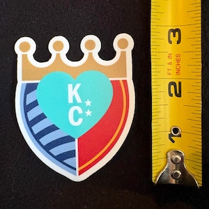 May include: A sticker with a shield design featuring a blue and white striped pattern, a red section, and a turquoise heart with the letters "K*C*" inside. The shield is topped with a gold crown.