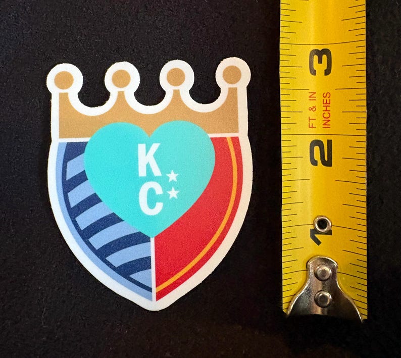 May include: A sticker with a shield design featuring a blue and white striped pattern, a red section, and a turquoise heart with the letters "K*C*" inside. The shield is topped with a gold crown.