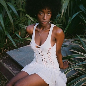 May include: A woman with an afro hairstyle wears a white, lace-trimmed, sleeveless dress with a cut-out design. The dress has a ruffled hem and is tied at the waist.