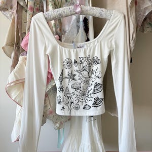 Puede incluir: Top blanco de manga larga con cuello cuadrado y un gráfico de conchas negras. También se ve una falda blanca con dobladillo escalonado y ribete de encaje. La ropa está colgada en una percha rosa con otras prendas.