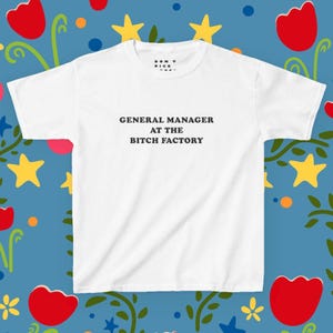 Può includere: T-shirt bianco con la scritta nera "GENERAL MANAGER AT THE BITCH FACTORY". La maglietta ha girocollo e maniche corte. Lo sfondo è un motivo blu con fiori rossi e stelle gialle.