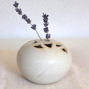 May include: A speckled, off-white ceramic vase with geometric cutouts, holding sprigs of dried lavender. The spherical vase has a textured surface and a neutral color palette, suitable for home decor.