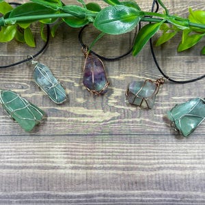 May include: Five gemstone pendants on black cords. The gemstones are various colours, including green and purple, and are wrapped in silver or copper wire. The pendants are displayed on a wooden surface with green foliage.
