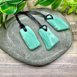 May include: Three rectangular, light green pendants with black cords rest on a grey stone. The pendants have a smooth, polished appearance. Green foliage is in the background, and the surface is a wooden texture.