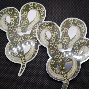 Peut inclure: Deux autocollants de serpent vert avec des fleurs blanches sur un fond noir. Les serpents sont enroulés en forme de cœur.