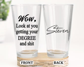 May include: Two clear glass pint glasses with black text. The front glass says "Wow, Look at you getting your DEGREE and shit". The back glass says "Steven".