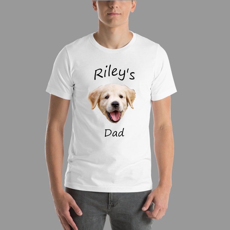 May include: White t-shirt with a golden retriever puppy's face and the text "Riley's Dad" printed on it.