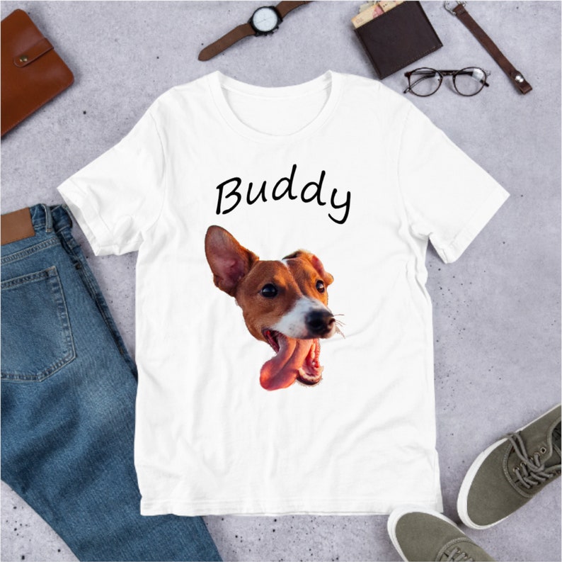 May include: White t-shirt with a brown and white dog's face printed on it. The dog is sticking its tongue out and the text "Buddy" is printed above the dog's face.