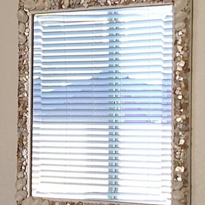 May include: A rectangular mirror with a frame decorated with seashells and small, iridescent pieces. The mirror reflects a window with horizontal blinds, creating a bright and airy feel. The frame's colors are neutral, with shades of white, beige, and brown.