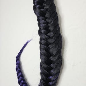 Puede incluir: Una extensión de cabello trenzado en un esquema de dos tonos. La parte superior es negra, que transiciona a un púrpura intenso en los extremos. La trenza es gruesa y parece estar hecha de fibras sintéticas.
