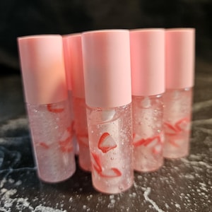 May include: A collection of six clear lip gloss tubes with pink caps. Each tube contains a clear liquid with small red strawberry slices. The lip gloss tubes are arranged closely together.