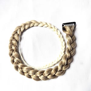 May include: A hair extension piece featuring a thick, braided strand of light brown and blonde hair. The braid is attached to a small, black metal snap clip at one end, allowing for easy attachment. The hair has a multi-tonal appearance.
