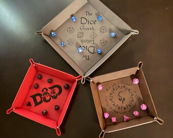 May include: Three leather dice trays with different designs. The top tray is brown with the text "The Dice Giverh Takerh Did The" and blue dice. The left tray is red with the text "D&D" and black dice. The right tray is brown with the text "Are you a Druid? Legally you have to tell me" and pink dice.
