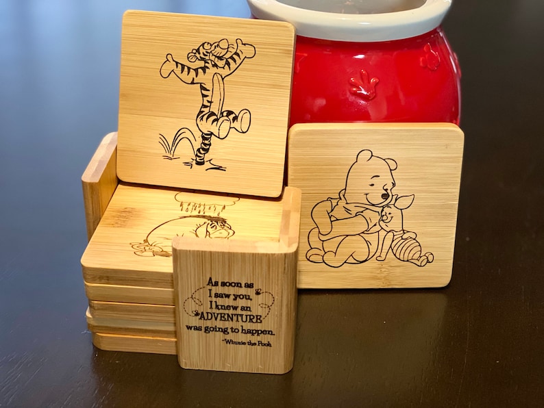 May include: Set of six wooden coasters featuring Winnie the Pooh and Tigger characters. The coasters are engraved with images of the characters and a quote from Winnie the Pooh: "As soon as I saw you, I knew an adventure was going to happen." - Winnie the Pooh