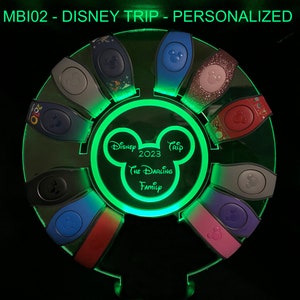 以下が含まれることがあります： ミッキーマウスのシルエットと「Disney Trip 2023 The Darling Family」というテキストが書かれた緑色のライトアップディスプレイ。ディスプレイには、10個のカラフルなディズニーマジックバンドの円形が置かれています。