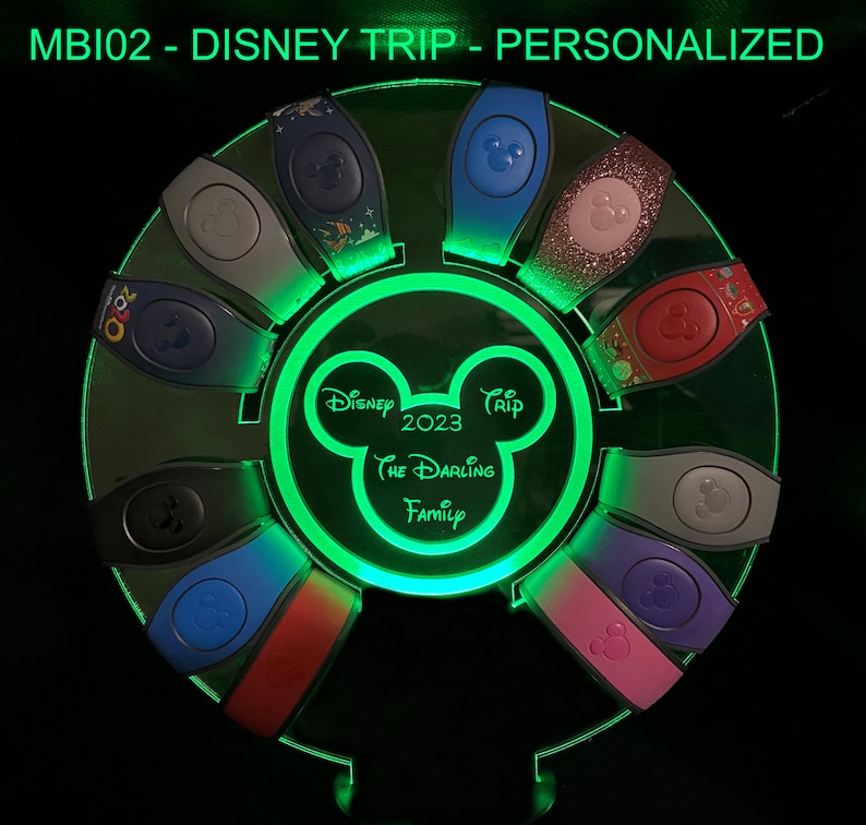 以下が含まれることがあります： ミッキーマウスのシルエットと「Disney Trip 2023 The Darling Family」というテキストが書かれた緑色のライトアップディスプレイ。ディスプレイには、10個のカラフルなディズニーマジックバンドの円形が置かれています。