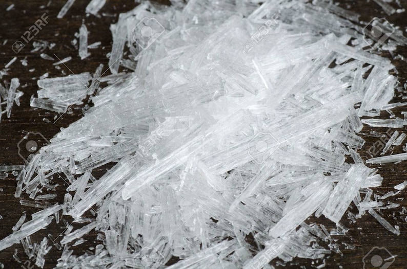 May include: A close-up of a pile of clear, crystalline shards on a dark brown surface. The shards are irregularly shaped and have a translucent appearance.