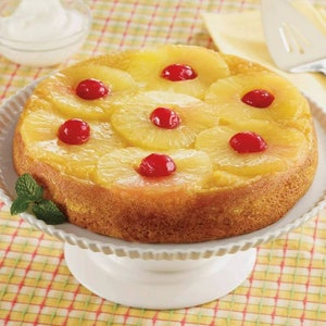 Può includere: Una torta rovesciata all'ananas gialla con ciliegie maraschino rosse in cima, su un supporto per torte bianco.